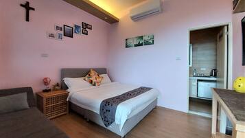 A bedroom with a bed, a sofa, a small table, a wall-mounted air conditioner, and a kitchen area visible through an open door.