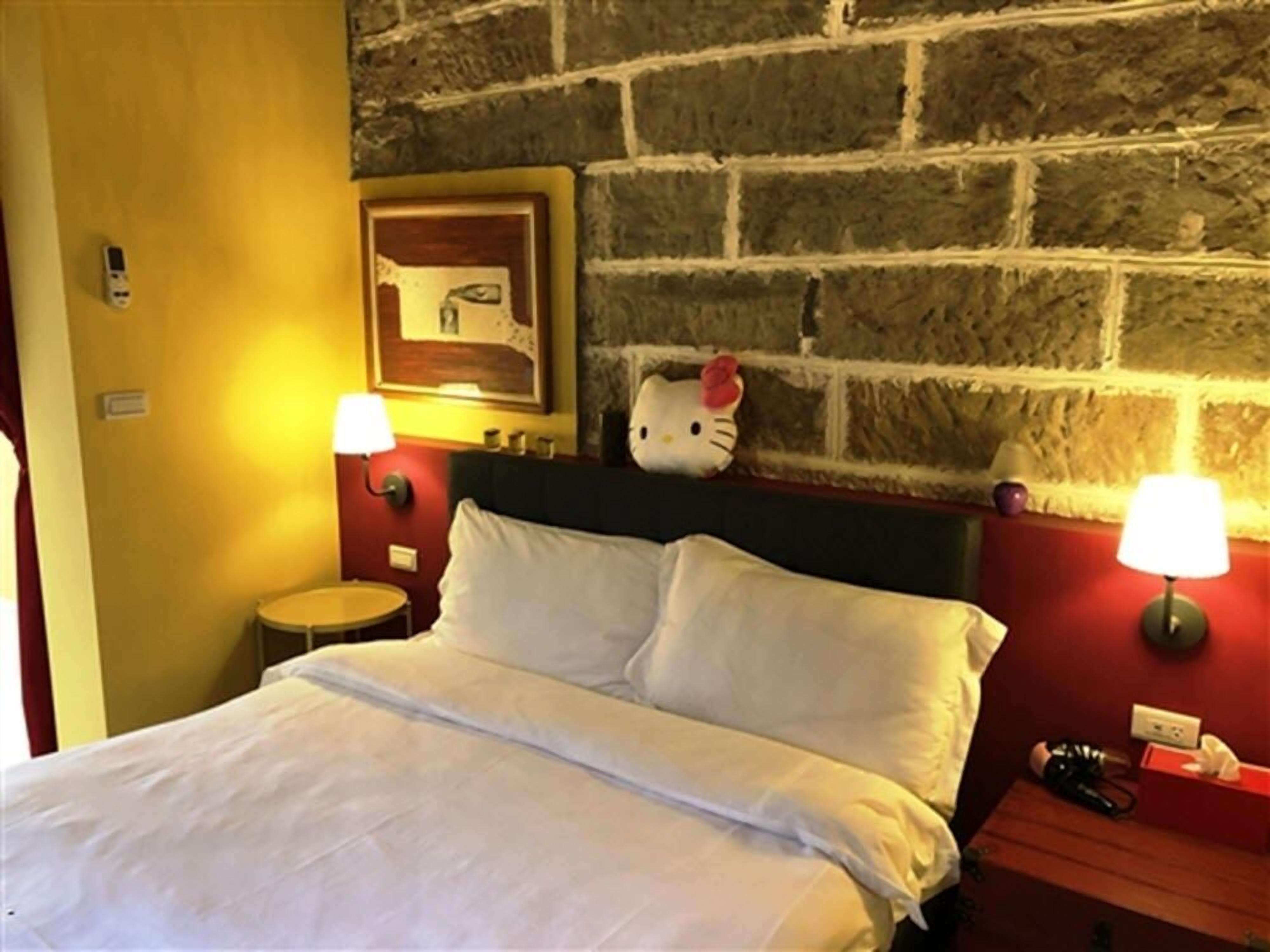 A hotel room with a brick wall, a bed with white linens, two bedside lamps, a Hello Kitty plush toy, and a small round table with a lamp.