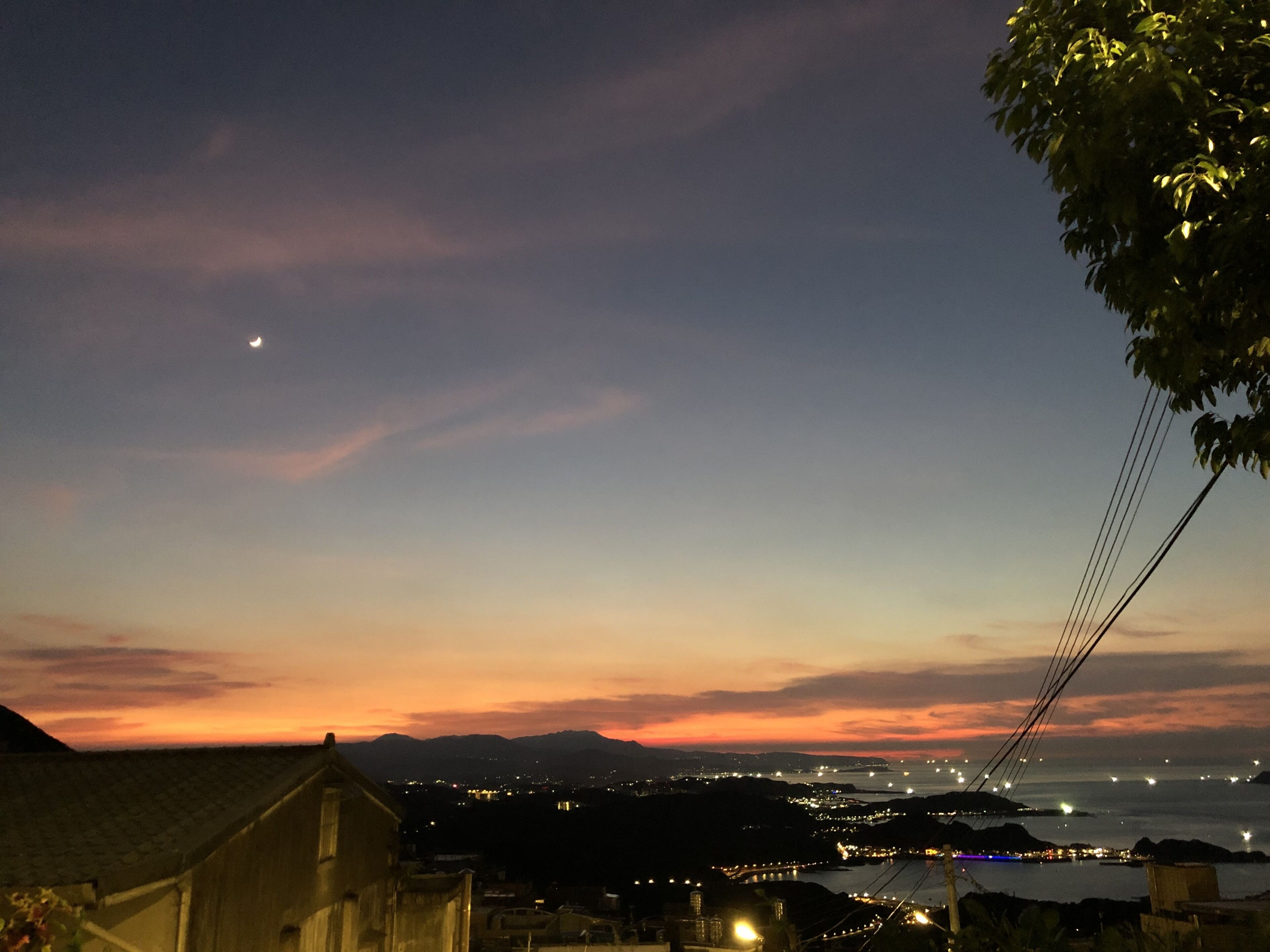 A nighttime view of a coastal city with a bridge, distant mountains, and a crescent moon.