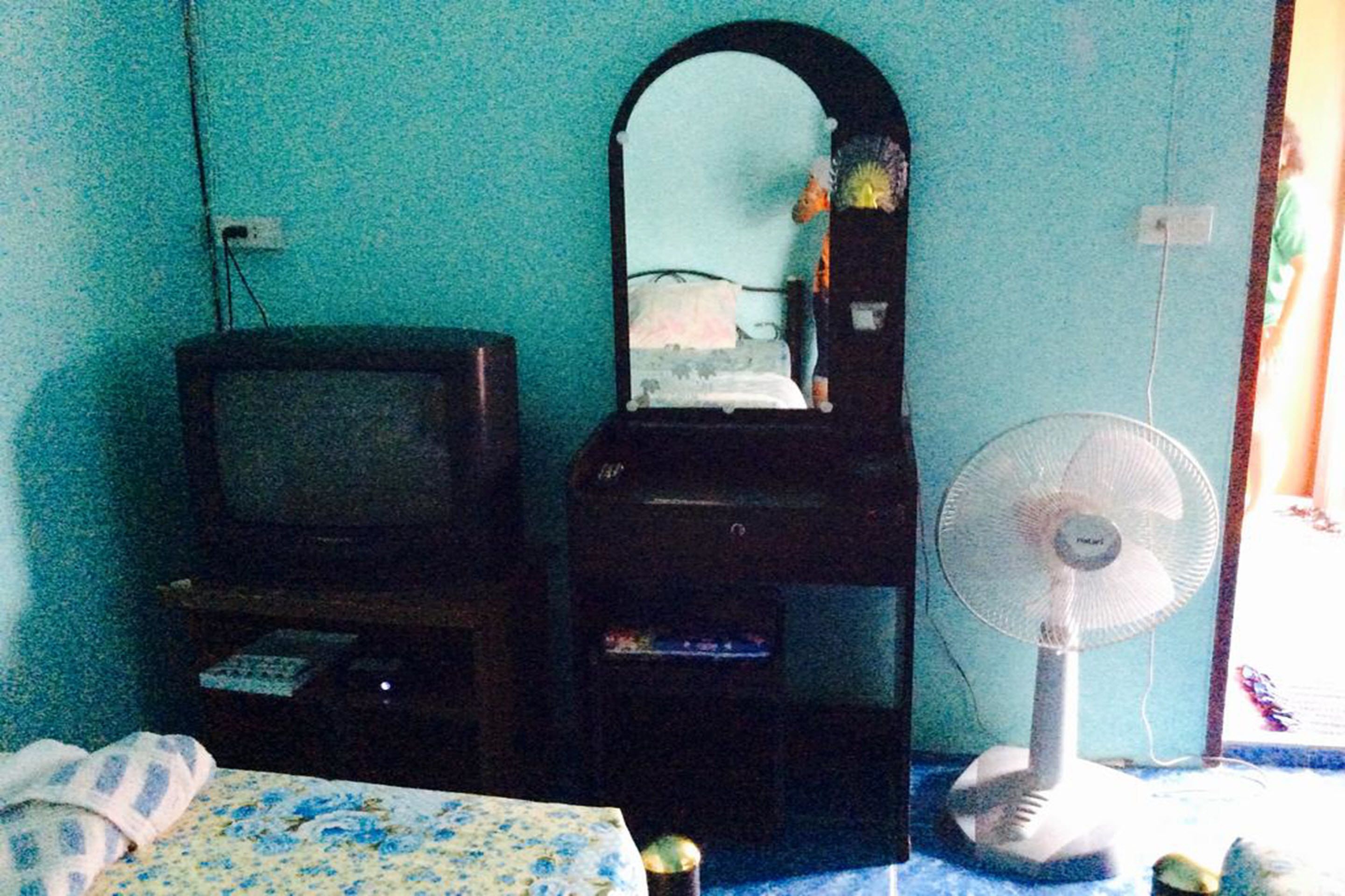 Una habitación con cama, televisión, espejo y ventilador de pie.