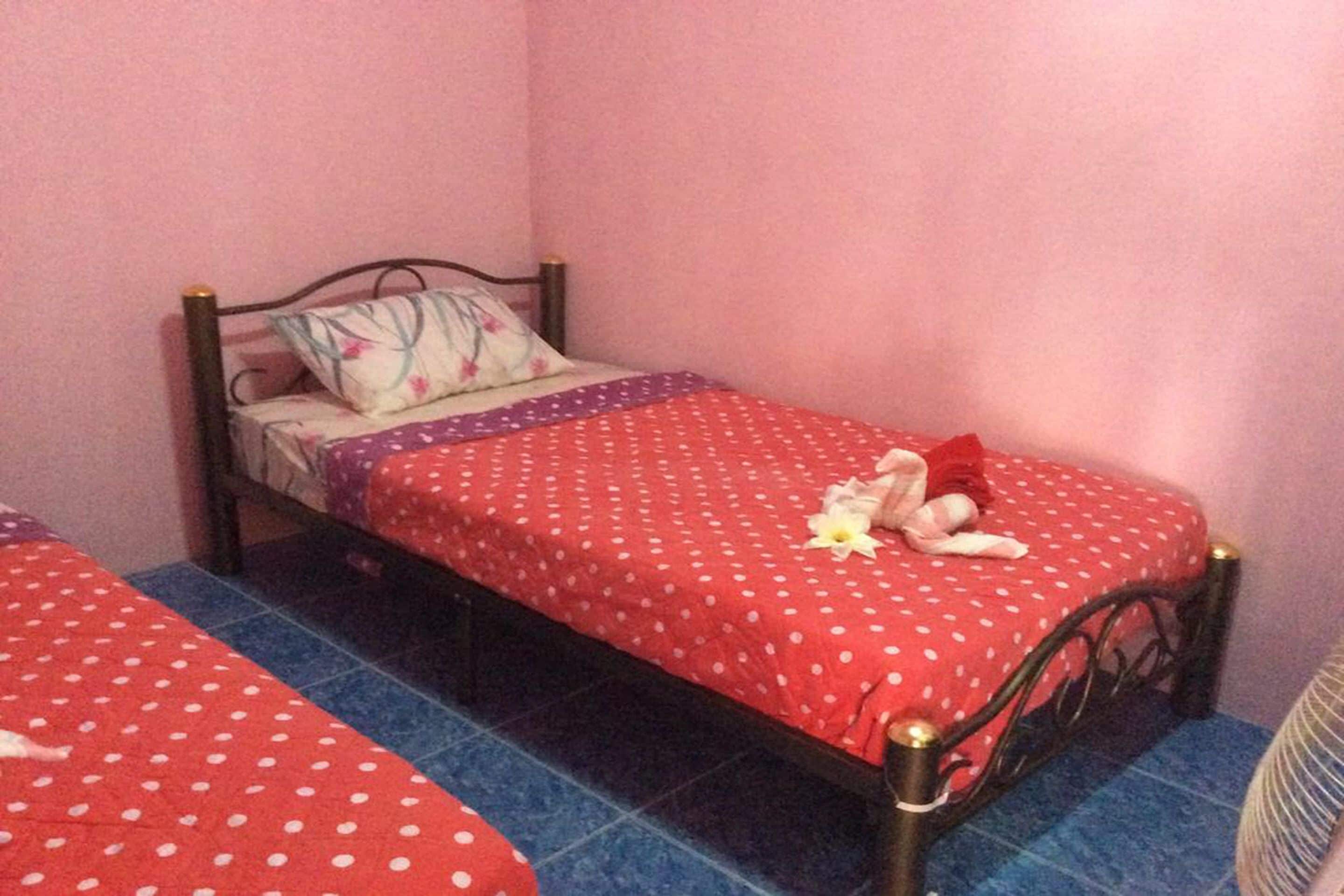 A single bed with a red polka dot bedspread and a floral pillow, placed on a blue-tiled floor in a room with pink walls.
