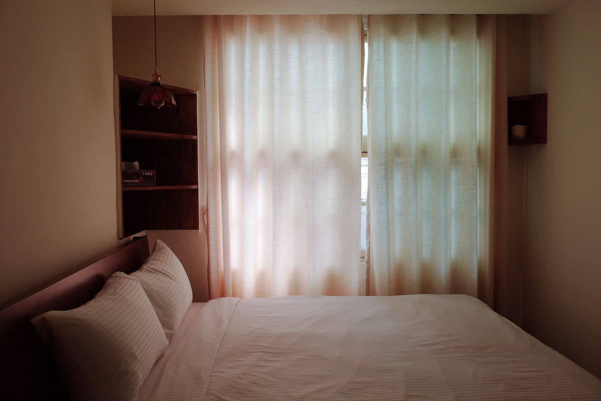 A hotel room with a bed, a shelf with items, and sheer curtains.