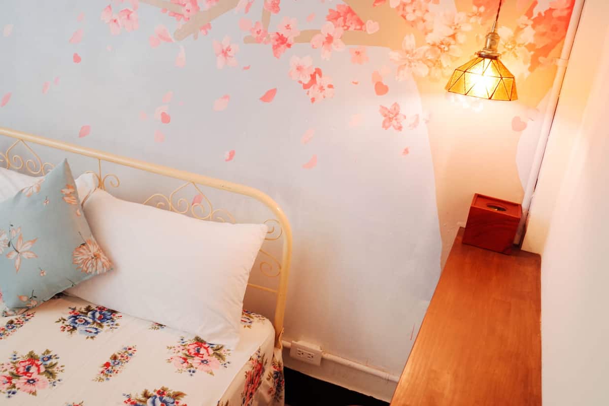 A bedroom with a bed, pillows, a wall with floral wallpaper, a hanging light fixture, and a wooden nightstand.