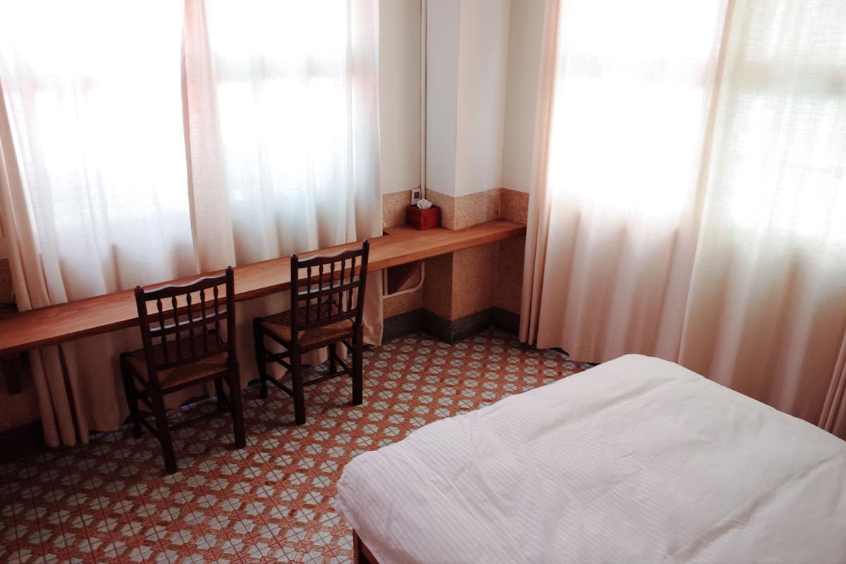 A room with a bed, a desk with two chairs, and a window with curtains.
