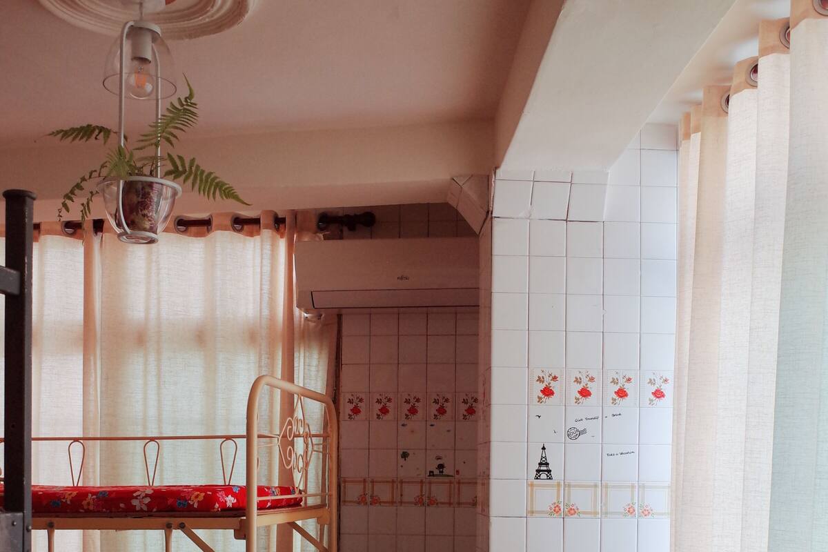 A bunk bed with a floral bedspread, a small table, a chair, a potted plant, and a tiled wall with decorative tiles.