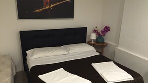 Un lit bien fait, avec du linge de lit blanc, une tête de lit sombre, une petite table de chevet ornée d’un vase de fleurs violettes et, au mur, un tableau représentant un pont.
