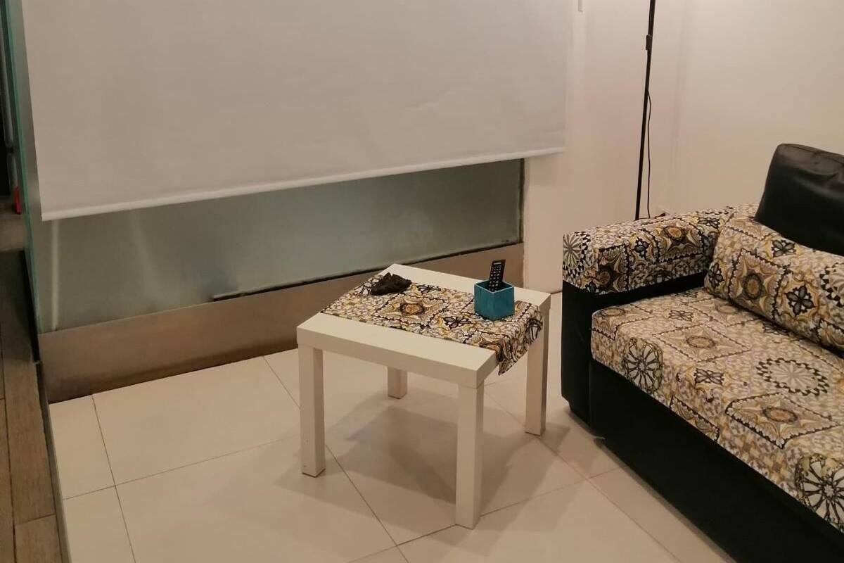 Une pièce avec un canapé blanc, un coussin décoratif à motifs, une petite table blanche avec une télécommande et une lampe sur pied.