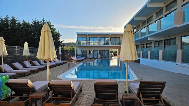 Zona de piscina con sillones reclinables y sombrillas, una terraza de madera y un edificio moderno con amplios ventanales.