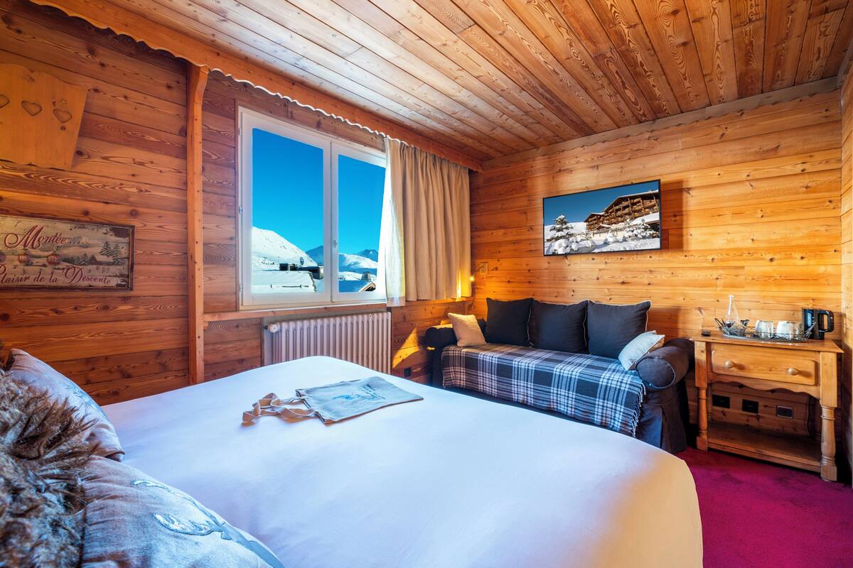 Une pièce de style cabane en bois, avec un lit, un canapé à carreaux, une table de chevet en bois et une fenêtre donnant sur des montagnes enneigées.