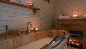 Una bañera de madera con ducha, un estante de madera con adornos, y una chimenea con leña.