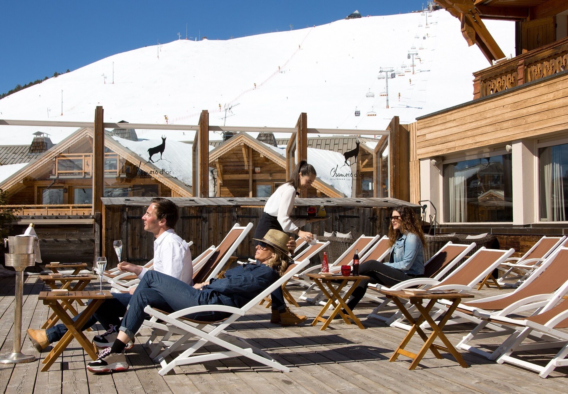 Une terrasse en bois avec des chaises longues et des tables, un paysage montagneux en toile de fond et une enseigne indiquant «Hôtel d’Or».