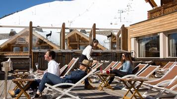Une terrasse en bois avec des chaises longues et des tables, un paysage montagneux en toile de fond et une enseigne indiquant «
Hôtel d’Or
».