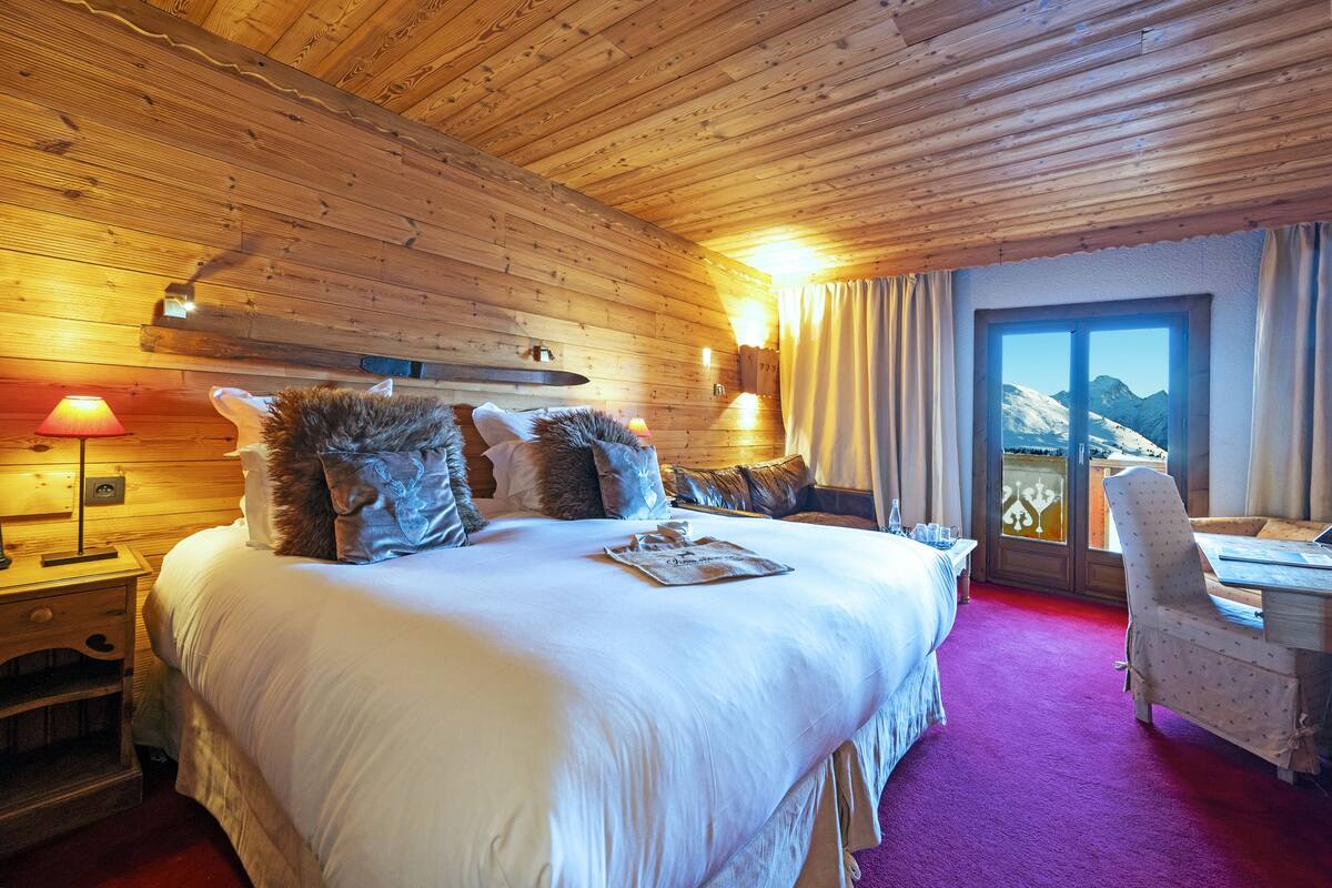Une chambre aux murs lambrissés, dotée d’un grand lit, d’un bureau et offrant une vue sur les montagnes grâce à une porte vitrée.