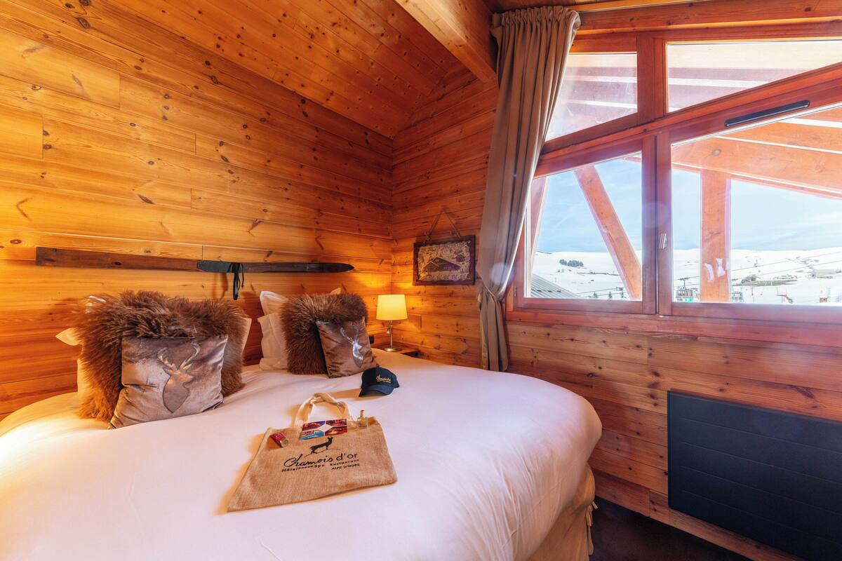 Une chambre dans un chalet en bois, avec un lit, des oreillers, un sac et une fenêtre donnant sur un paysage agréable.