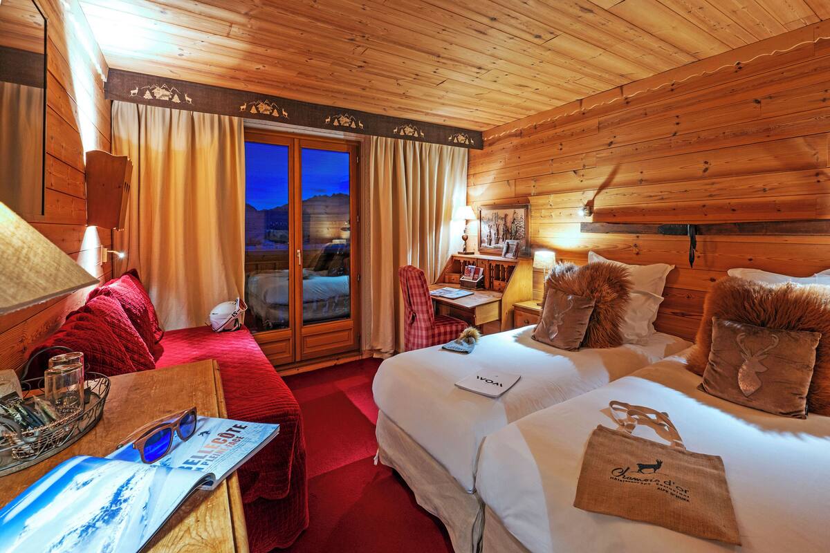 Une chambre d’hôtel chaleureuse avec des murs et un plafond en bois, un grand lit recouvert d’une couette blanche, un bureau avec une chaise et une vue sur les montagnes par la porte vitrée.