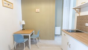 Ein kleiner, gut beleuchteter Raum mit einem Holztisch und zwei Stühlen, einem weißen Heizkörper sowie einer Küchenzeile mit Arbeitsplatte und Schränken.