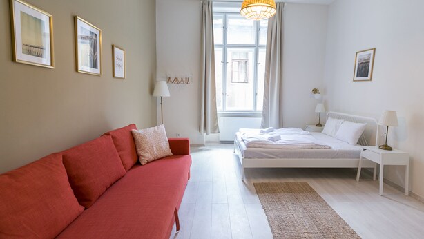 Ein modernes Schlafzimmer mit einem roten Sofa, einem weißen Bett, einem großen Fenster mit Vorhängen und gerahmten Bildern an der Wand.
