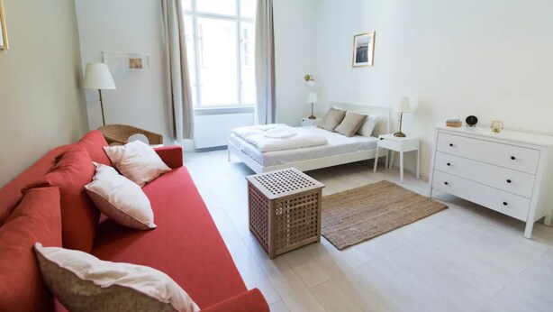 Un dormitorio moderno con un sofá rojo, una cama blanca, un taburete de madera y un aparador blanco.
