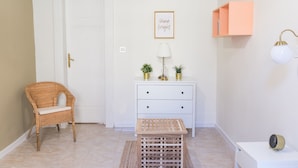 Una habitación con una silla de mimbre, un tocador blanco, un taburete de madera, una cita motivacional en un marco, una estantería montada en la pared y una mesita de noche con una lámpara.