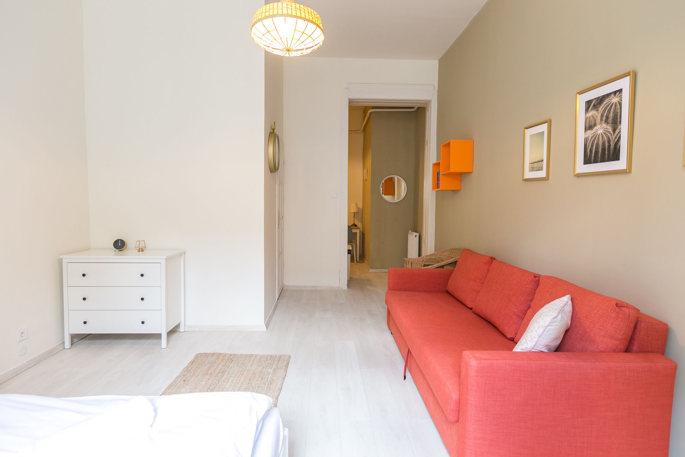 A bedroom with a bed, a white dresser, a red sofa, and a hallway leading to another room.