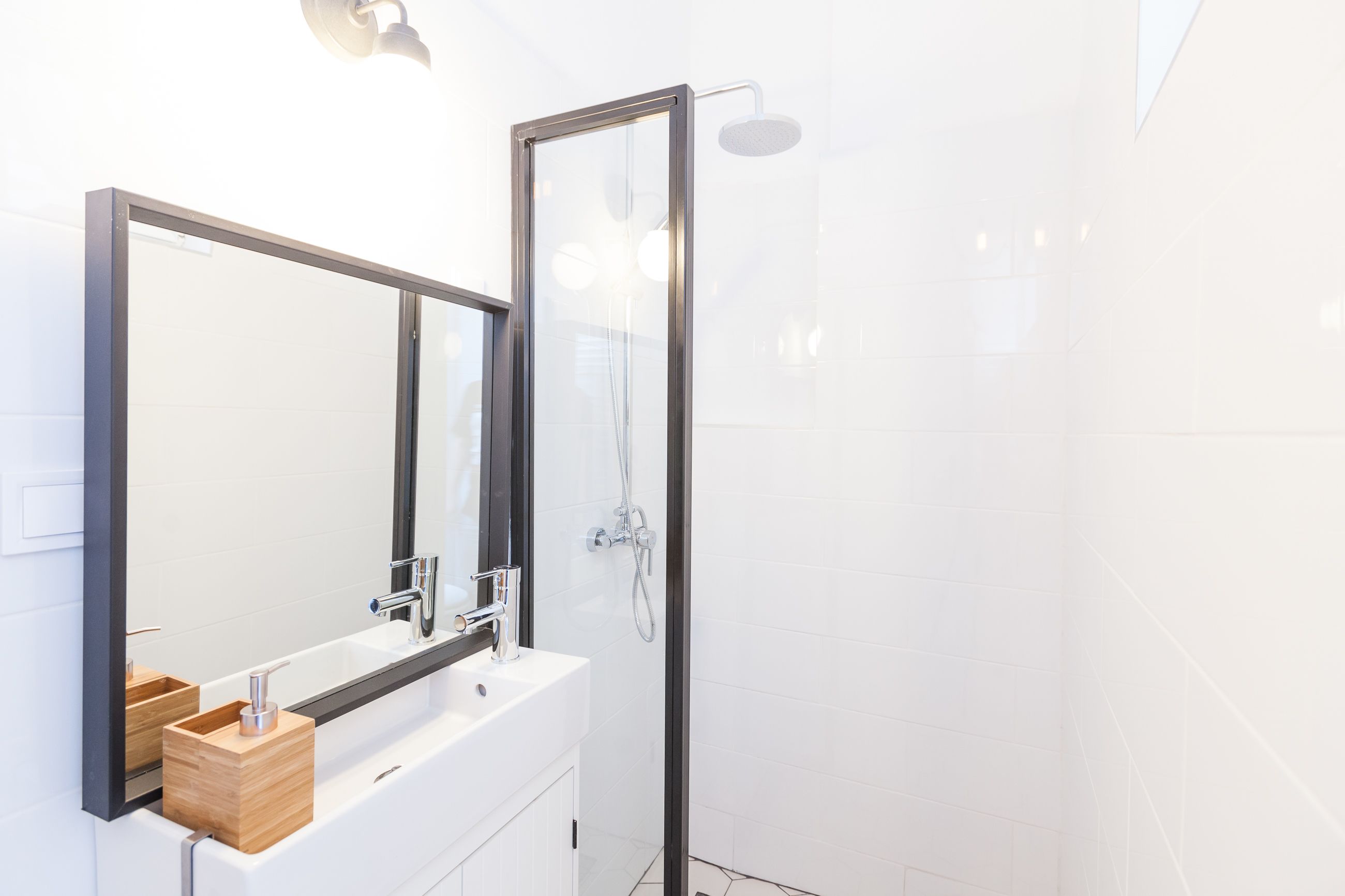 Een moderne badkamer met een douchecabine van glas, een grote spiegel en een wit wastafelmeubel met een houten zeepdispenser.