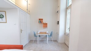 Una habitación pequeña y luminosa con una cama, una mesa con sillas, un estante pequeño con libros y una ventana.
