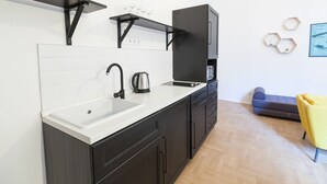 Cocina moderna con encimera blanca, armarios negros y horno integrado. Hay una cafetera en la encimera y una pequeña zona de estar con sofá y una silla amarilla.