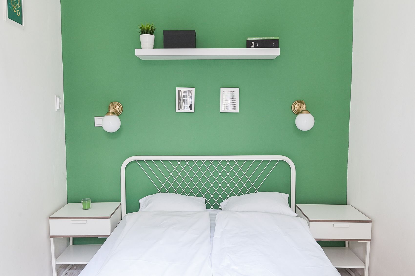 A bed with a white metal headboard, two bedside tables, a green wall, a shelf with a plant and books, and two wall-mounted lamps.