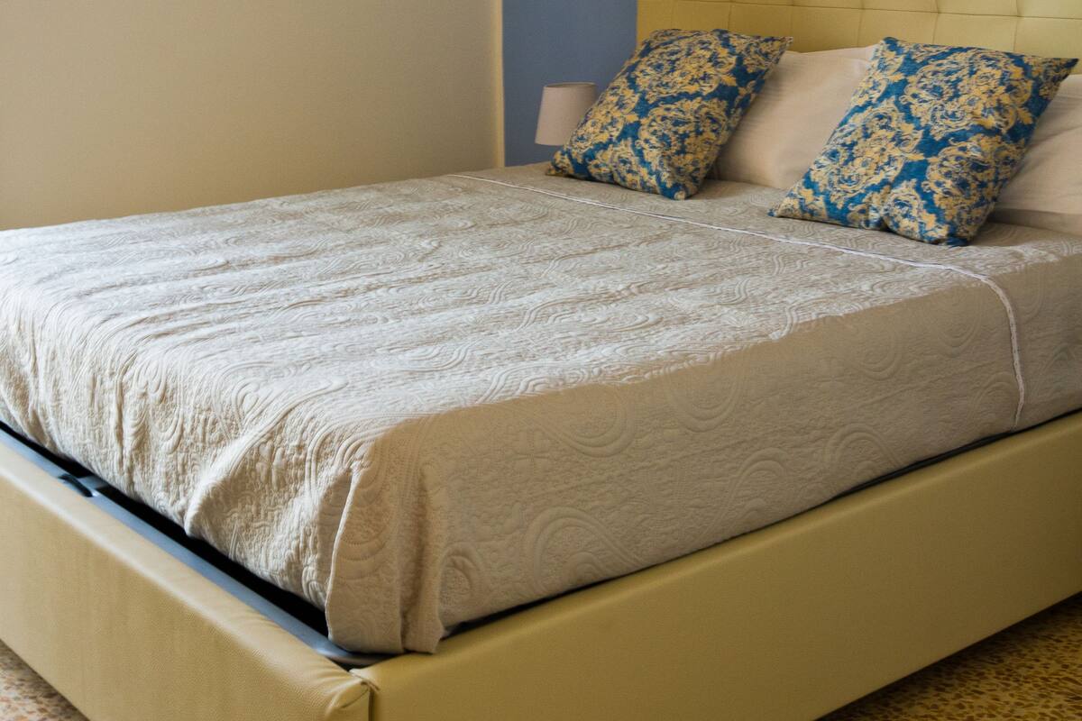 Un lit avec une tête de lit de couleur beige, deux coussins à motifs et un mur uni agrémenté d’un petit luminaire.