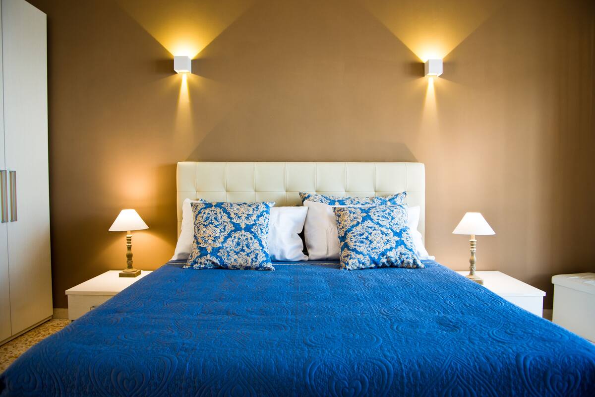 Un lit bien fait, recouvert d’une couvre-lit bleue et agrémenté d’oreillers à motifs, avec deux lampes de chevet placées de part et d’autre sur des tables de chevet blanches.