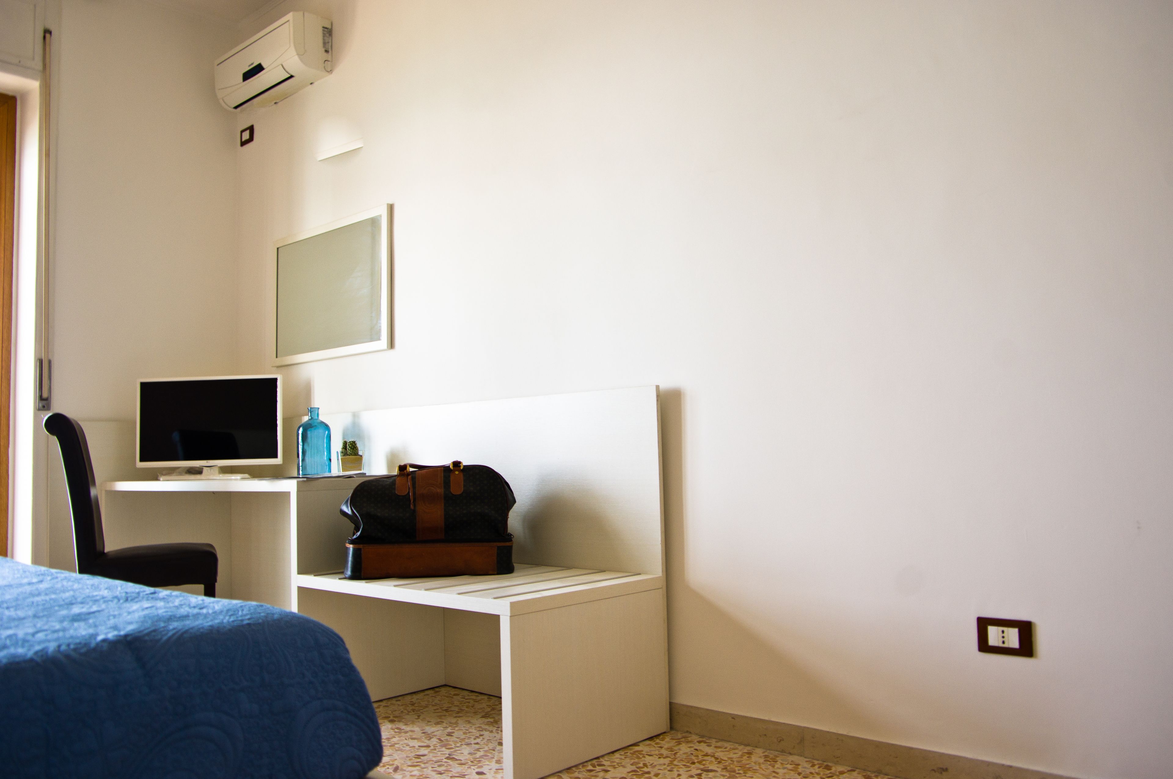 Une chambre d’hôtel comprenant un lit, un bureau avec un ordinateur et une chaise, une petite table avec un sac, et un climatiseur fixé au mur.