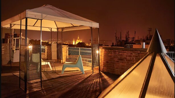 Un área de estar al aire libre con un toldo, una silla colgante y una barandilla con vistas a un paisaje urbano de noche.