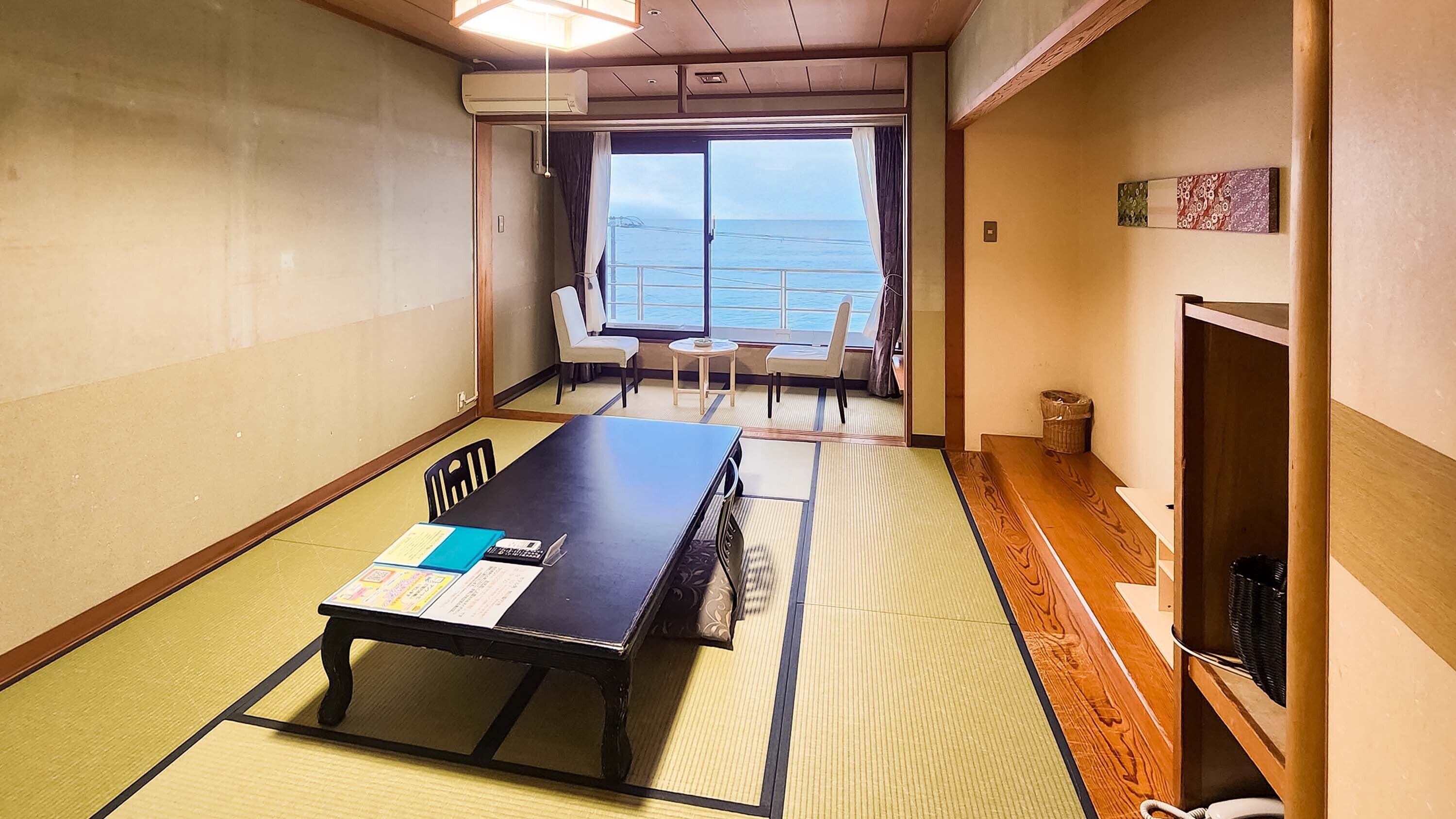 A room with tatami flooring, a low table with a menu, a chair, a small table with a cup, and a view of the sea through a large window.