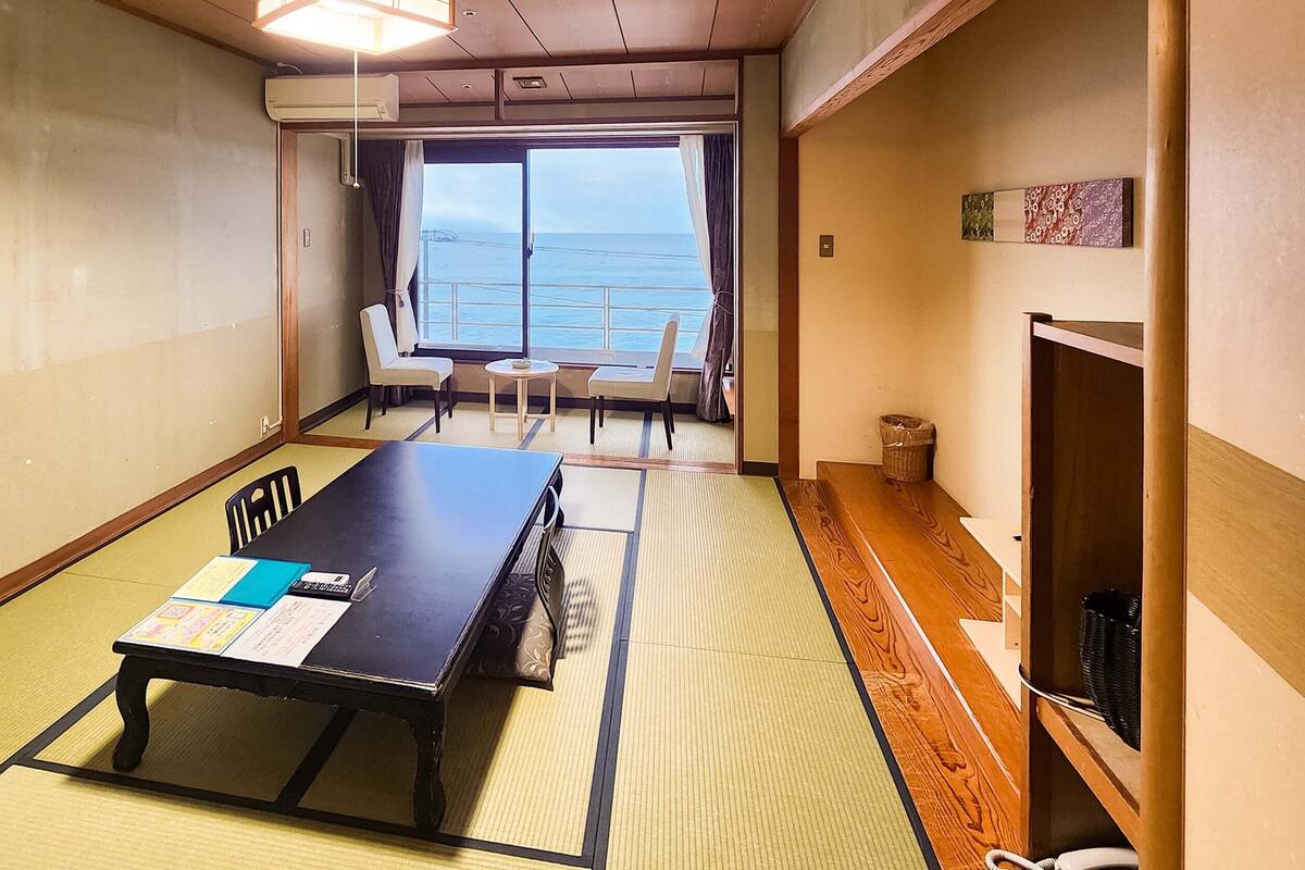 A room with tatami flooring, a low table with a menu, a chair, a small table with a cup, and a view of the sea through a large window.