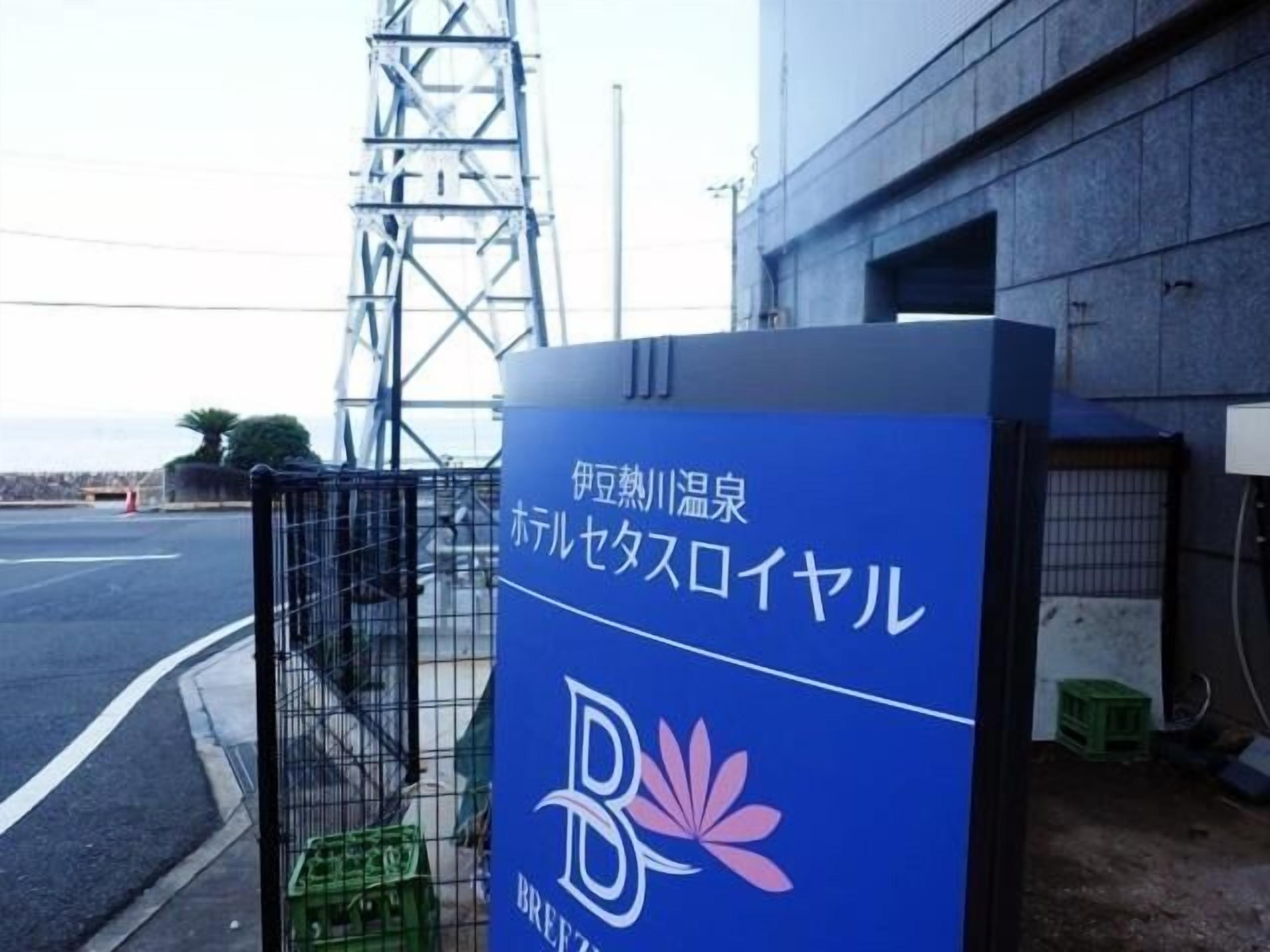 Un letrero azul con texto japonés y un logotipo, sujeto a una estructura metálica cerca de un edificio.
