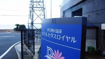 Un letrero azul con texto japonés y un logotipo, sujeto a una estructura metálica cerca de un edificio.