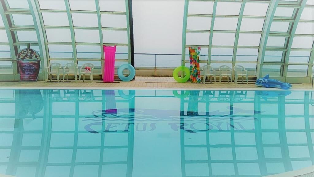 Una piscina interior con paredes de vidrio transparente, suelo de baldosas y varias sillas junto a la piscina.