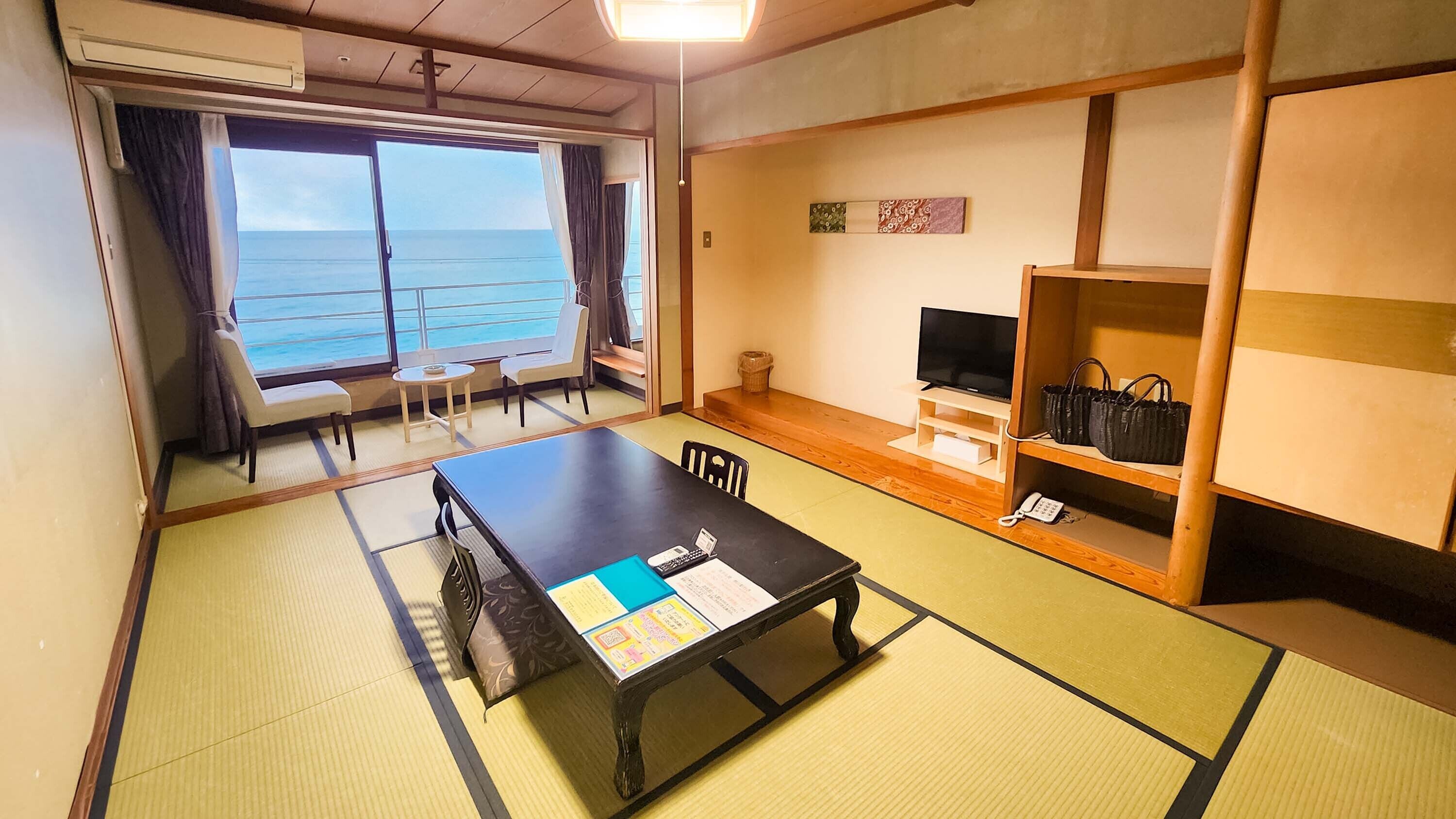 A room with tatami flooring, a table with papers, a TV, a shelf with bags, and a view of the sea.