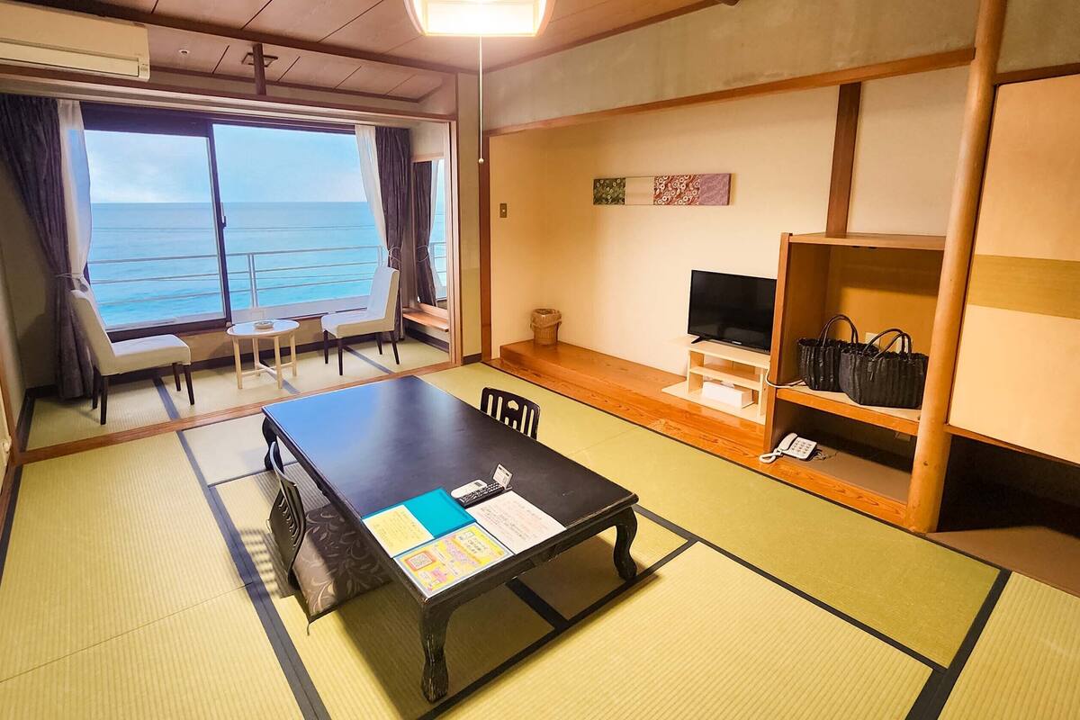 A room with tatami flooring, a table with papers, a TV, a shelf with bags, and a view of the sea.