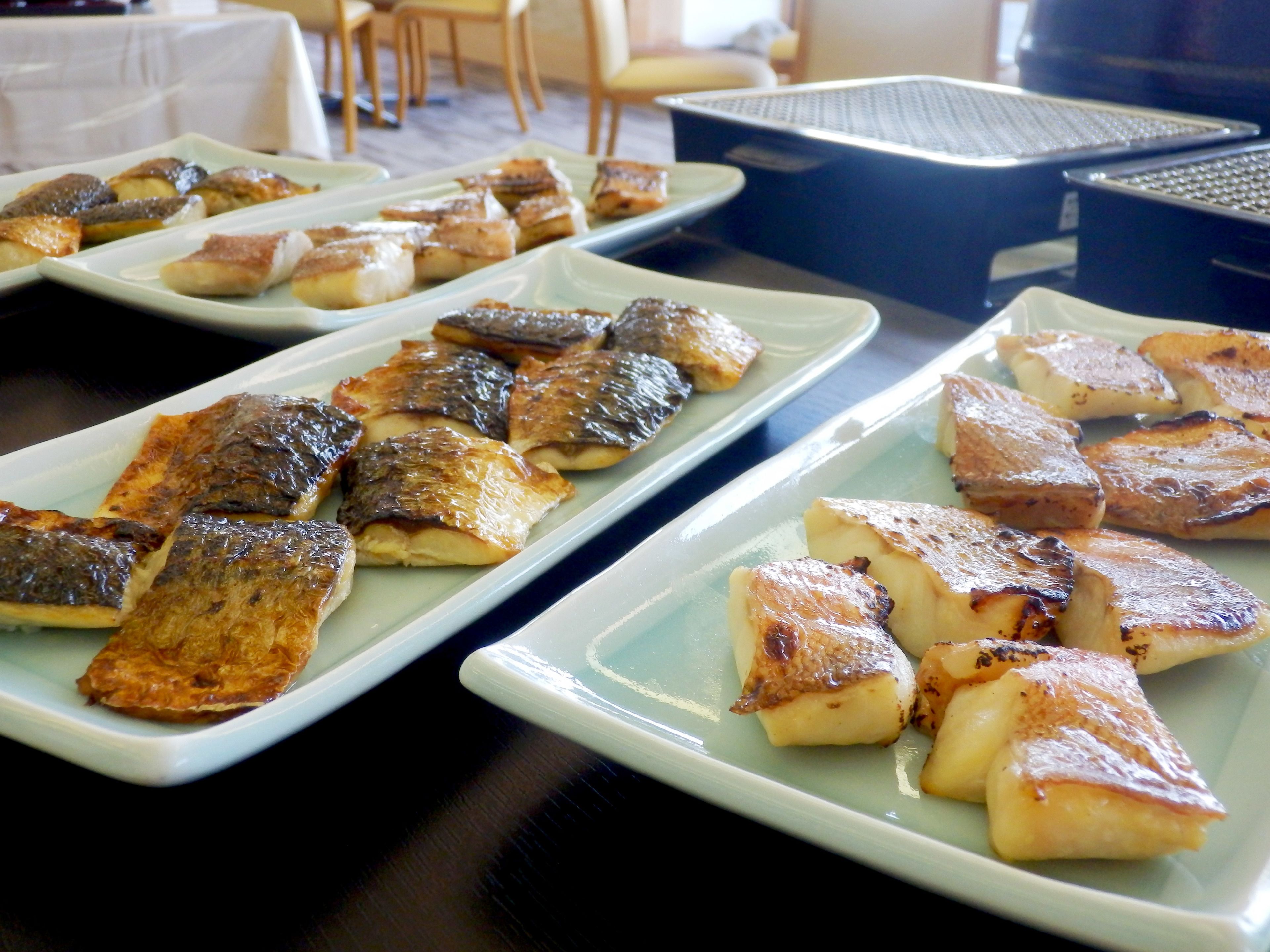 A buffet table with various dishes, including grilled fish and potatoes, arranged on rectangular plates.