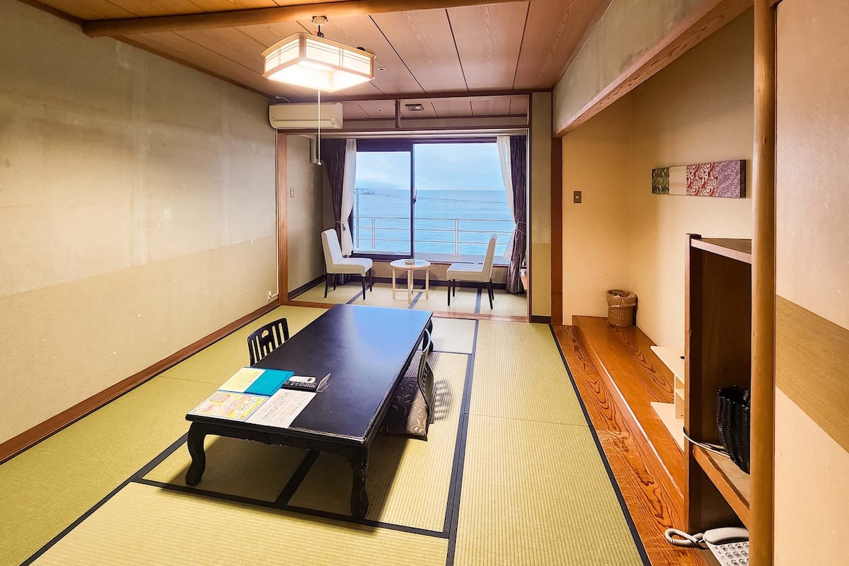 A room with tatami mats, a wooden table with papers, a chair, a small table with a cup, a window with a view of the sea, and a telephone.