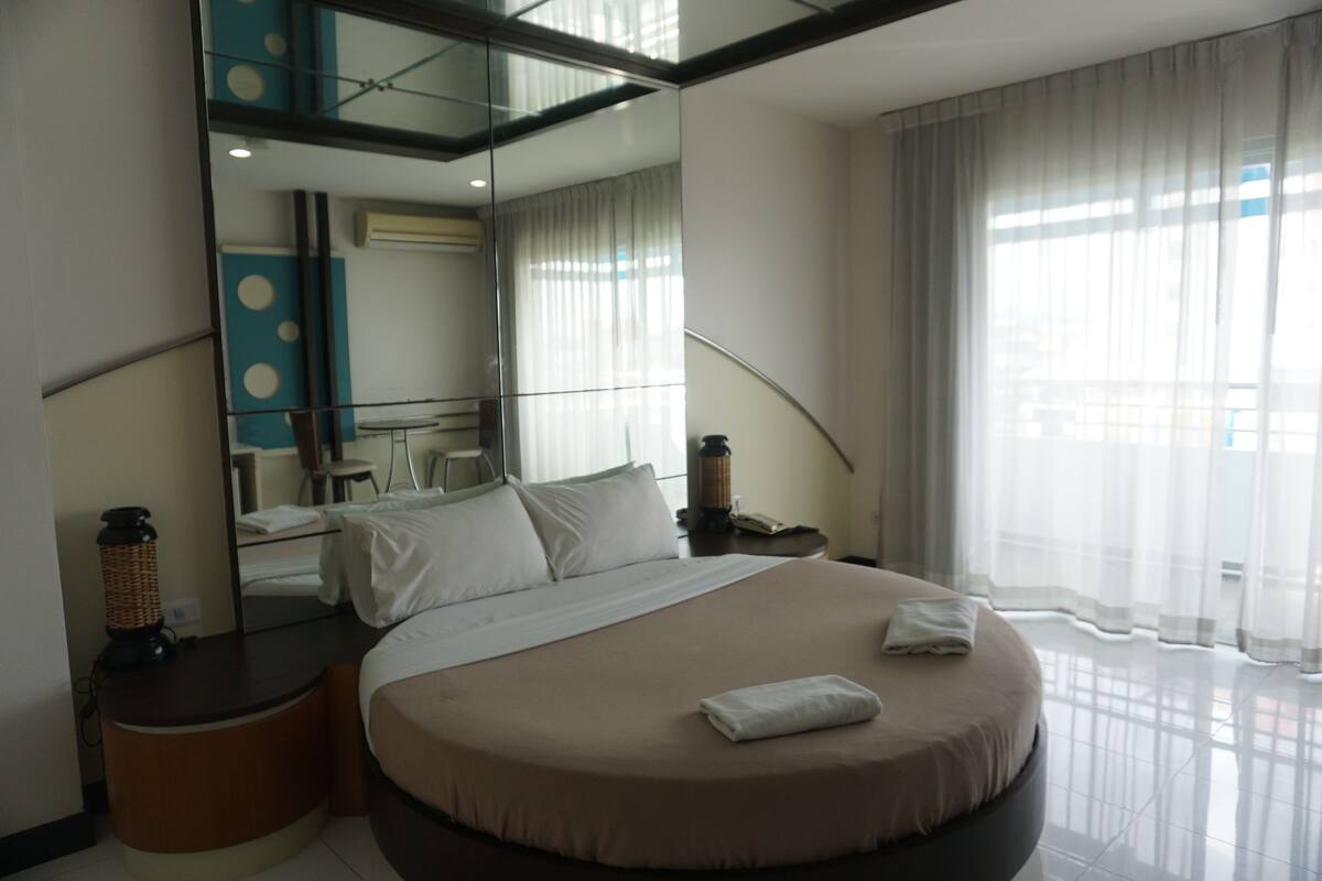 Ein modernes Hotelzimmer mit einem großen Bett, einem runden Nachttisch, einem Spiegel und Blick auf die Stadt durch transparente Vorhänge.