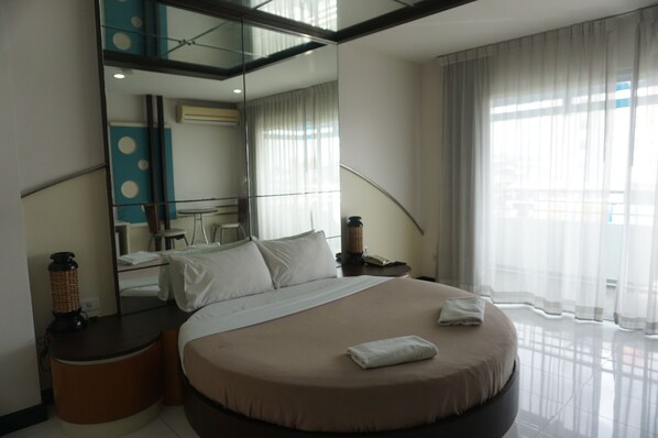 A modern hotel room with a large bed, a round bedside table, a mirror, and a view of the city through sheer curtains.