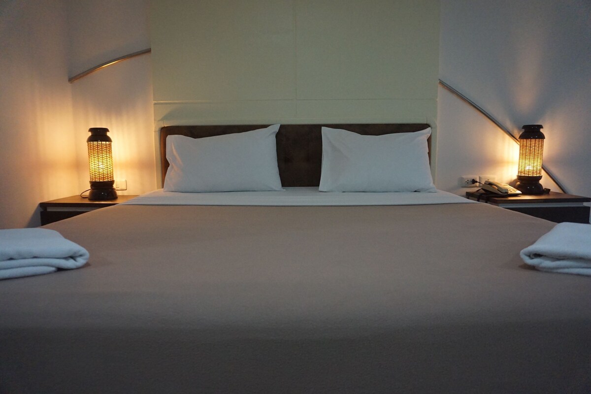 Een net opgemaakt bed met witte lakens, twee bedlampjes en een nachtkastje met een telefoon en een afstandsbediening.