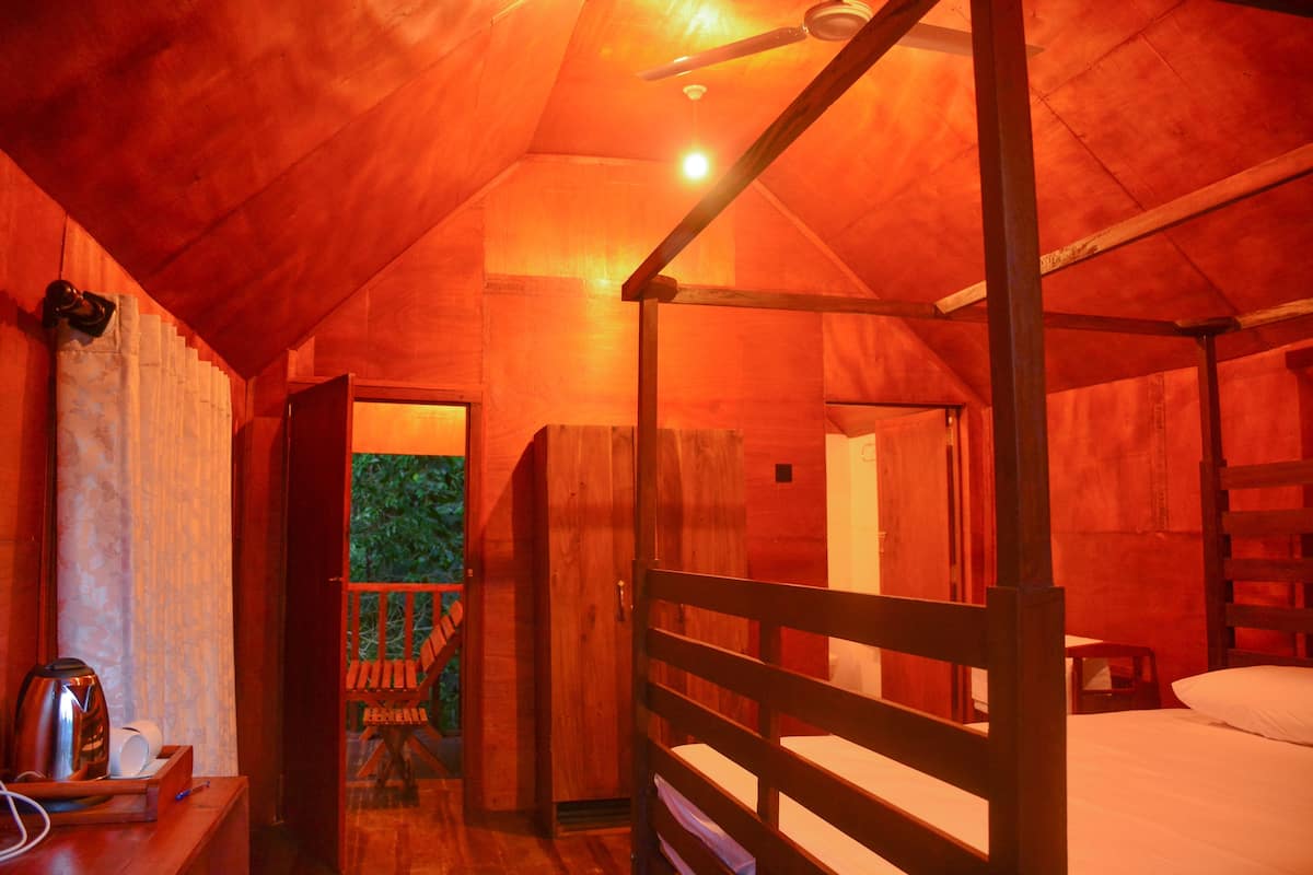 A cozy, single-room cabin with a wooden ceiling, a bed, a small table with a kettle, and a chair.