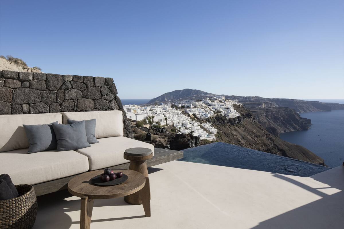 Una terraza con pared de piedra, un sofá, una mesa redonda de madera y vistas a un pueblo costero de edificios blancos.