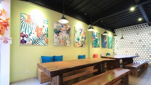Un restaurante con largas mesas y bancos de madera, colorida decoración mural tropical y lámparas colgantes.