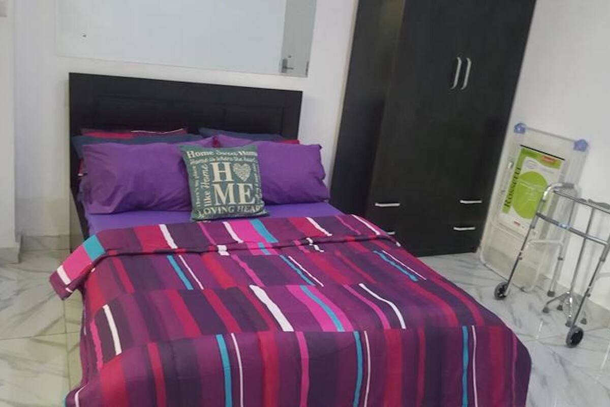 A bedroom with a bed, purple pillows, a striped blanket, a dark headboard, a wardrobe, and a cart with bottles.
