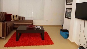 A living room with a sofa, a coffee table, a red rug, a TV, and a wall-mounted air conditioner. - Capital & County Apartment Ikate (Lekki)