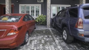 A parking area with two cars, a red sedan and a blue SUV, parked in front of a building with a grey facade and a white door. - Capital & County Apartment Ikate (Lekki)